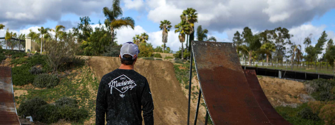 A person wearing a cap and a shirt with "Mayaries" text on the back stands in front of two metal ramps surrounded by trees and a cloudy sky.
