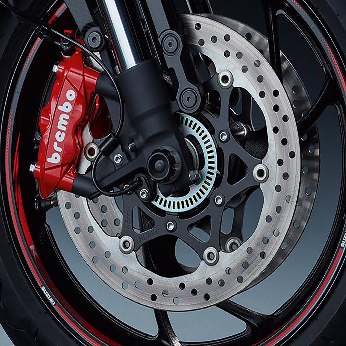 Close-up of a motorcycle front wheel showcasing a Brembo red brake caliper and a drilled disc brake. The tire has a red and white striped detail on the rim, and the suspension fork is visible.