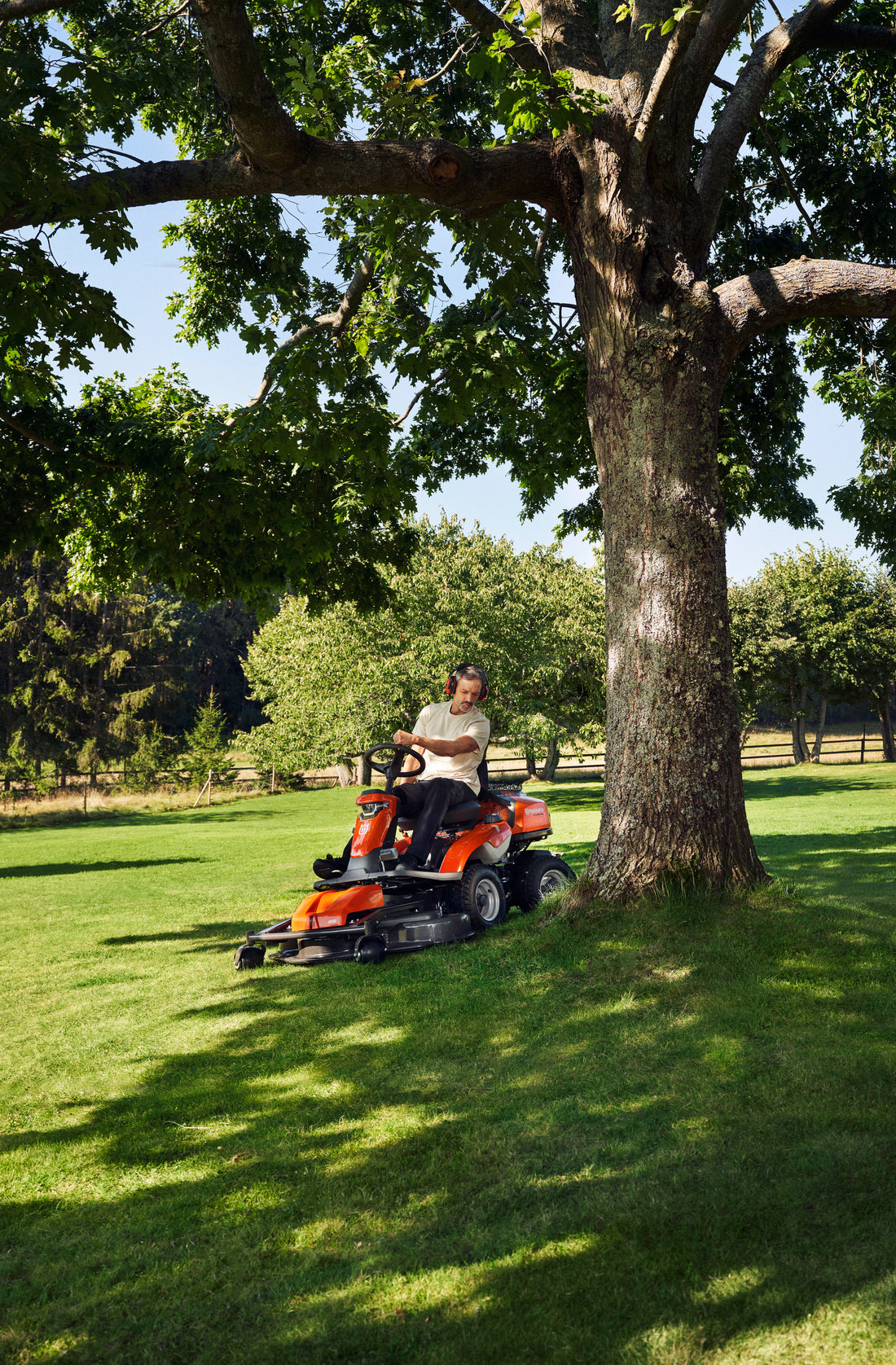 Person riding an orange lawn mower under a large tree in a sunny, green field. Trees and wooden fence are in the background.