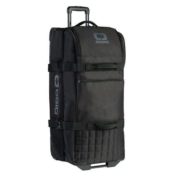 The Ogio TRUCKER Gear Bag - Black by Ogio is a rectangular rolling duffel bag featuring a padded handle on top, separate compartments, and a retractable handle. The bag displays dark grey OGIO logos on the front and sides, has two compression straps around the middle, and is made with eco-friendly PVC construction.