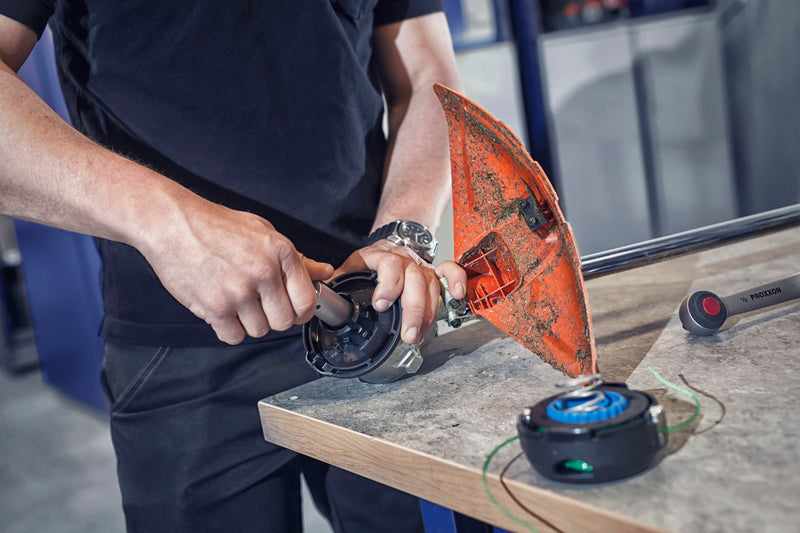 A person in a black shirt is disassembling an orange trimmer head in a workshop. Nearby, a new black and blue trimmer head and a tool stamped with "P. Precision" lies on a workbench. The person is using both hands to work on the equipment.