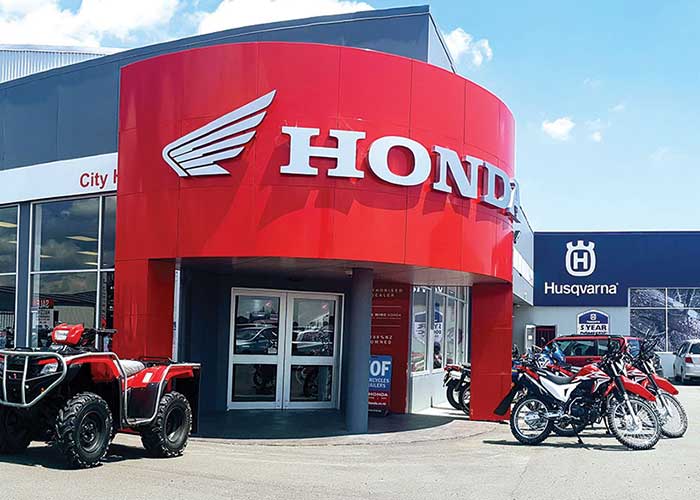 A Honda dealership with a red, curved entrance showcasing the Honda logo and winged emblem. Several Honda motorcycles and an ATV are parked in front. Adjacent is a Husqvarna sign on a nearby building against a blue sky background.