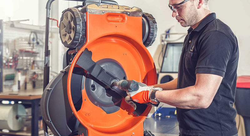 A technician wearing safety glasses and gloves uses a power tool to adjust the blade of an orange lawn mower flipped upside down in a workshop. Various tools and equipment are visible in the background.