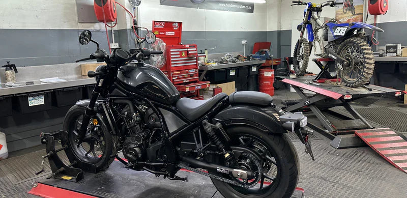 A motorcycle repair shop with a black cruiser motorcycle and a dirt bike on lifts. The room has toolboxes, shelves with equipment, and a gray and red color scheme. A banner is visible on the wall.