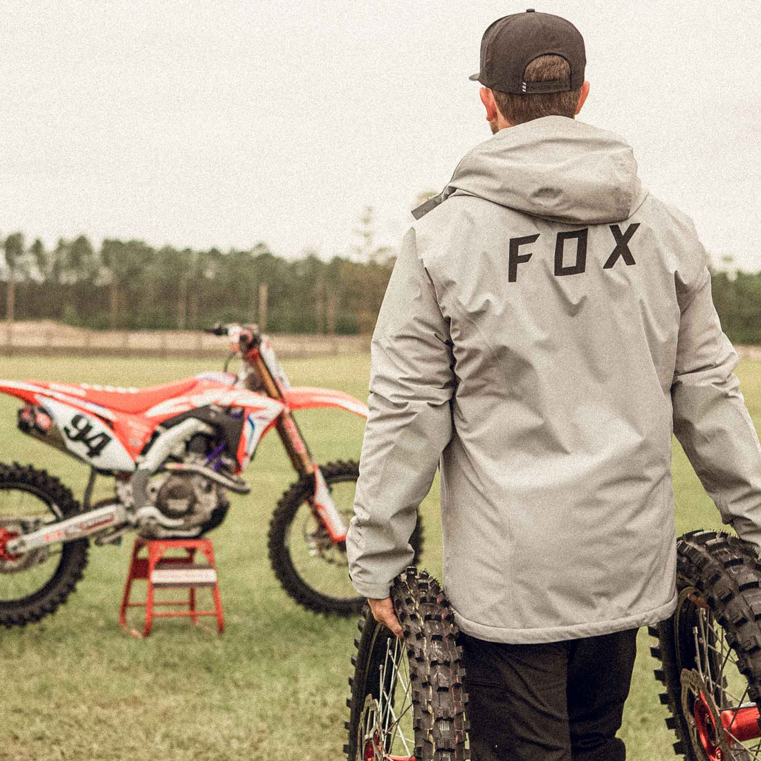 Guy at a motocross track holding on to a set of dirt bike wheels, wearing a Fox Racing Jacket. 