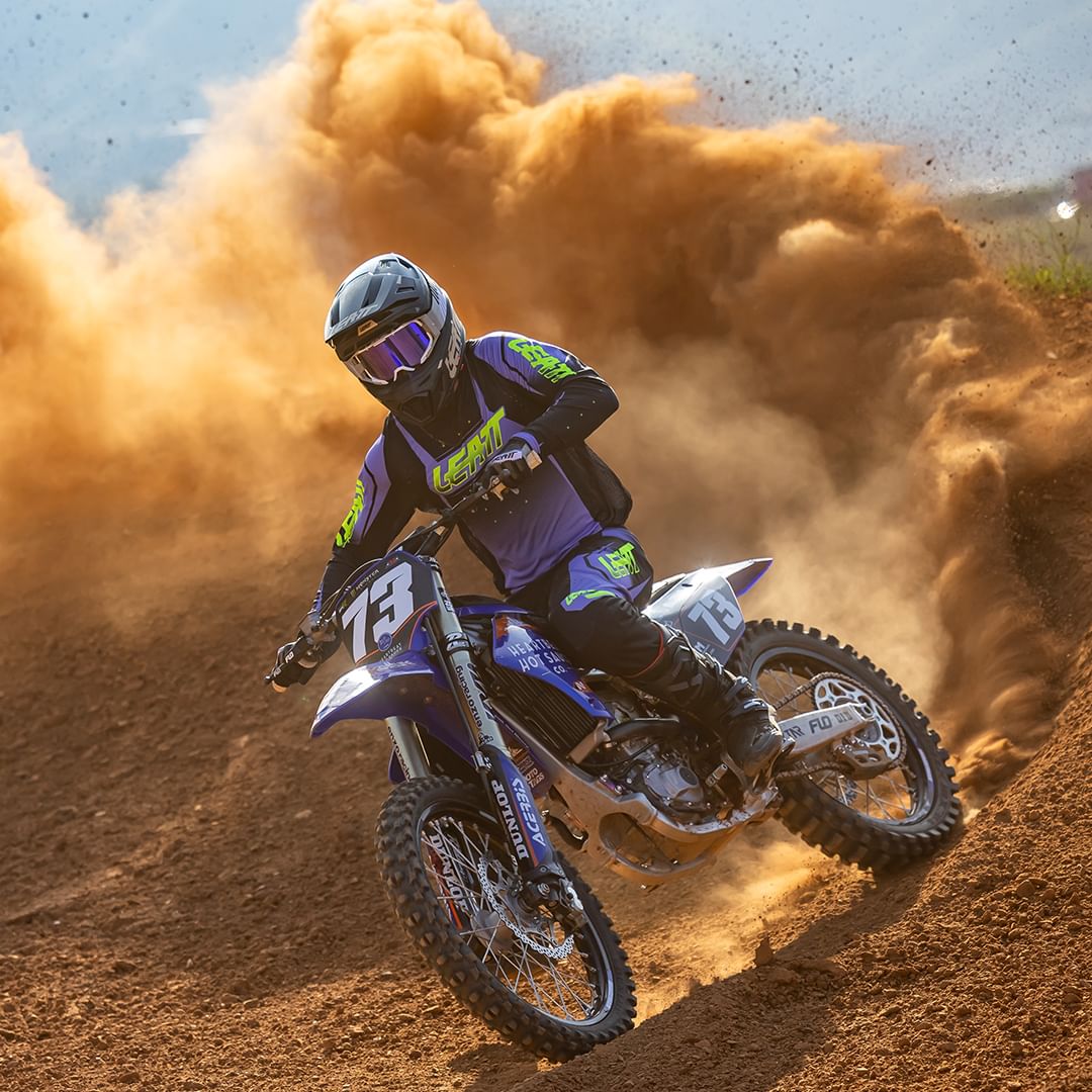 A motocross rider wearing a purple and black leatt MX gear with the number 13 on the bike and suit rides through a dirt track, kicking up a large cloud of dust behind them. They wear a helmet with a reflective visor.