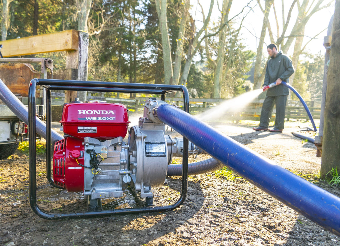 A person in outdoor workwear uses a blue hose connected to a red Honda WB20XT water pump to spray water on a dirt surface. The pump is placed on the ground in a sunlit area with trees and a wooden fence in the background.