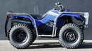 The Ventura Yamaha YFM 350FA Grizzly 4x4 Auto (12-17) ATV features a blue frame with large off-road tires, a tow ball, and metal racks on both ends. It sits on a concrete surface against a black wall and includes a black seat with mud flaps for utility and style.