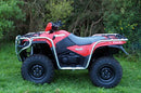 The Ventura Suzuki ATV LT-A 500 (13-18) in red, equipped with bull-bars, large black tires, and silver racks at both ends, is parked on the grass amidst green bushes and trees. It features a custom company logo on its side.