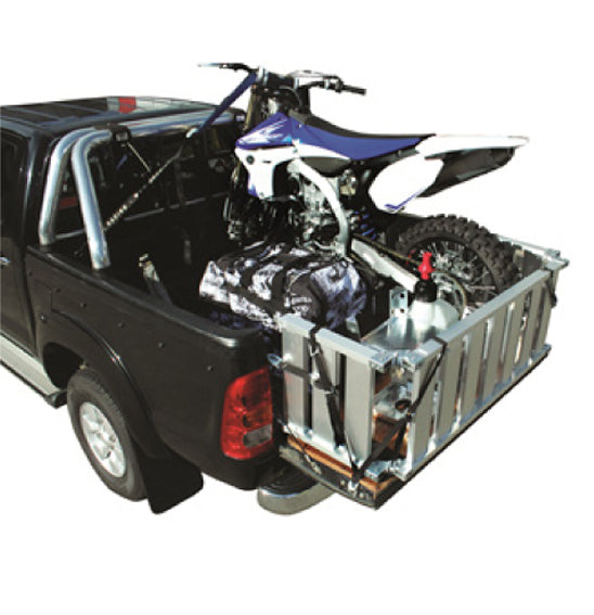 An X-TECH pickup truck with its tailgate down carries a motorcycle in the truck bed. The motorcycle is secured along with a camouflage bag and a metal container. The lightweight aluminum X-TECH Ute Bed Extender Ramp on the tailgate is presumably for loading and unloading the motorcycle.