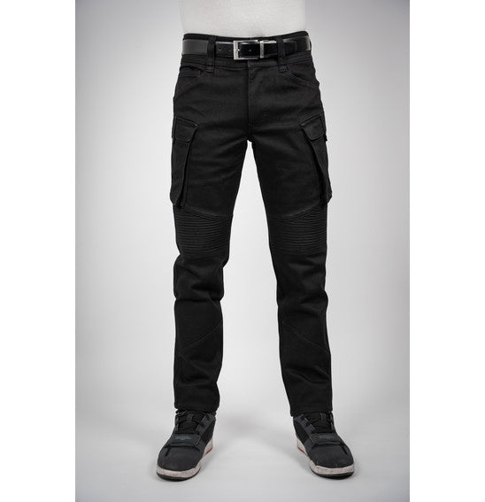 A person wearing Bull-It's jet black Jackal Motorcycle Jeans in a combat style, paired with a black belt and black sneakers with white soles, stands facing away. The jeans feature detailed stitching and zipper accents. The background is plain light gray.