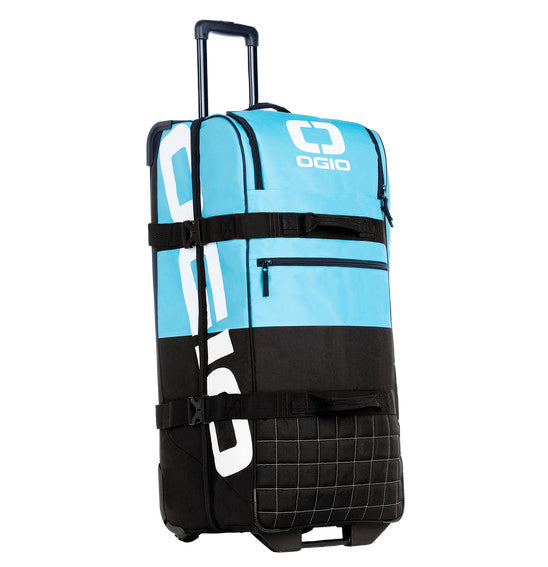 The OGIO TRUCKER Gear Bag - Maui is a large, wheeled travel bag with an extending handle that stands upright. Predominantly blue with black accents and white detailing, this eco-friendly PVC gear bag features four large compartments and multiple straps for securing items.