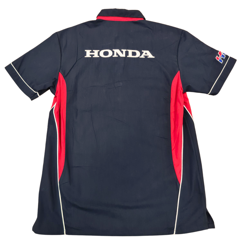 The HRC Honda HRC Pit Crew Shirt is a short-sleeved, button-up shirt featuring a stylish navy blue and red color scheme. It includes the Honda logo in white on the left chest area, two front pockets with red flaps, and a small flag patch on the right sleeve, offering a blend of performance and casual comfort for true fans.