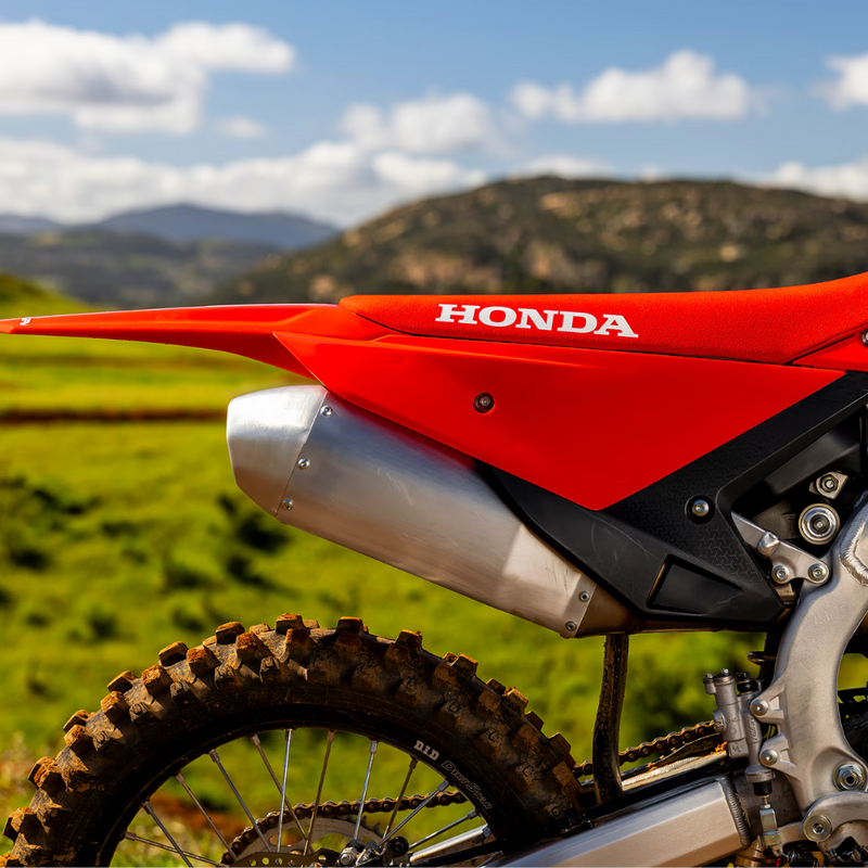 A side profile view of the 2025 Honda CRF450R showcases a striking red dirt bike with black tires and silver rims against a white background. The bike features a sleek frame, a visible engine, and prominent Honda branding on the seat and front forks, embodying its Supercross championship heritage.