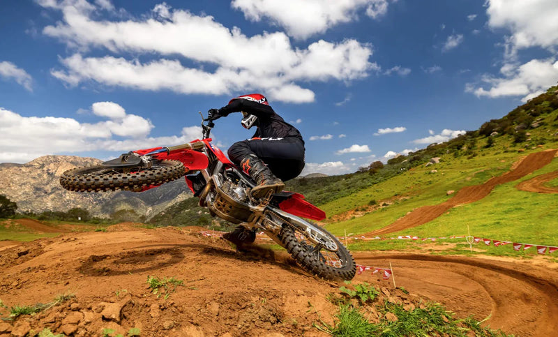 Side view of a red 2025 Honda CRF250R motocross dirt bike with black tires, visible spokes, and a silver exhaust pipe. The bike features the Honda logo on the seat and a number graphic on the side panel. Equipped with HRC Launch Control and a sturdy frame, it’s designed for optimal off-road use.