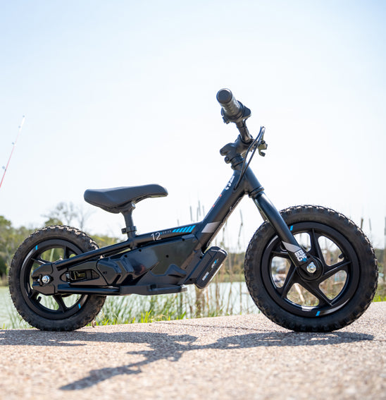 At a dirt track event, children lined up on STACYC 12eDRIVE electric balance bikes with 12” composite wheels, wearing colorful helmets and gear. A crowd watches in the background, and flags hang above them, capturing the vibrant energy of the scene.