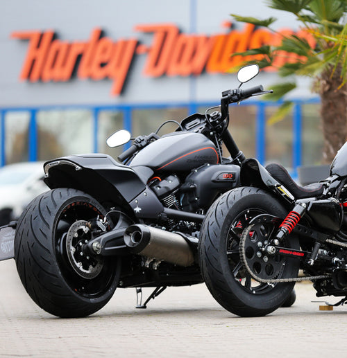 Two black Harley-Davidson motorcycles, sporting robust Metzeler CRUISETEC - Performance Cruiser tires by METZELER, are parked in front of a Harley-Davidson dealership. The image showcases the motorcycles' large rear tires, sleek design, and a hint of urban surroundings with the dealership's branded sign in the background.