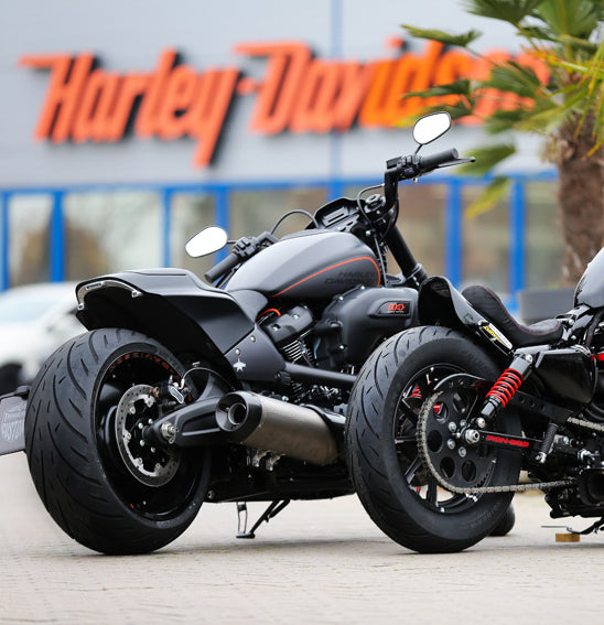 Two black Harley-Davidson motorcycles, sporting robust Metzeler CRUISETEC - Performance Cruiser tires by METZELER, are parked in front of a Harley-Davidson dealership. The image showcases the motorcycles' large rear tires, sleek design, and a hint of urban surroundings with the dealership's branded sign in the background.