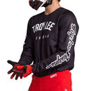 A person wearing the GP PRO JERSEY BOLTZ BLACK by Troy Lee Designs, featuring "TROY LEE DESIGNS" printed in white on the chest and sleeves, paired with red pants, red and black gloves, and a matching helmet. They are holding their hands out in front.