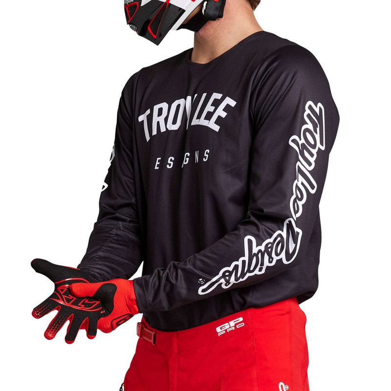 A person wearing the GP PRO JERSEY BOLTZ BLACK by Troy Lee Designs, featuring "TROY LEE DESIGNS" printed in white on the chest and sleeves, paired with red pants, red and black gloves, and a matching helmet. They are holding their hands out in front.