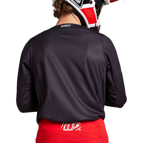 A person wearing the GP PRO JERSEY BOLTZ BLACK by Troy Lee Designs, featuring "TROY LEE DESIGNS" printed in white on the chest and sleeves, paired with red pants, red and black gloves, and a matching helmet. They are holding their hands out in front.