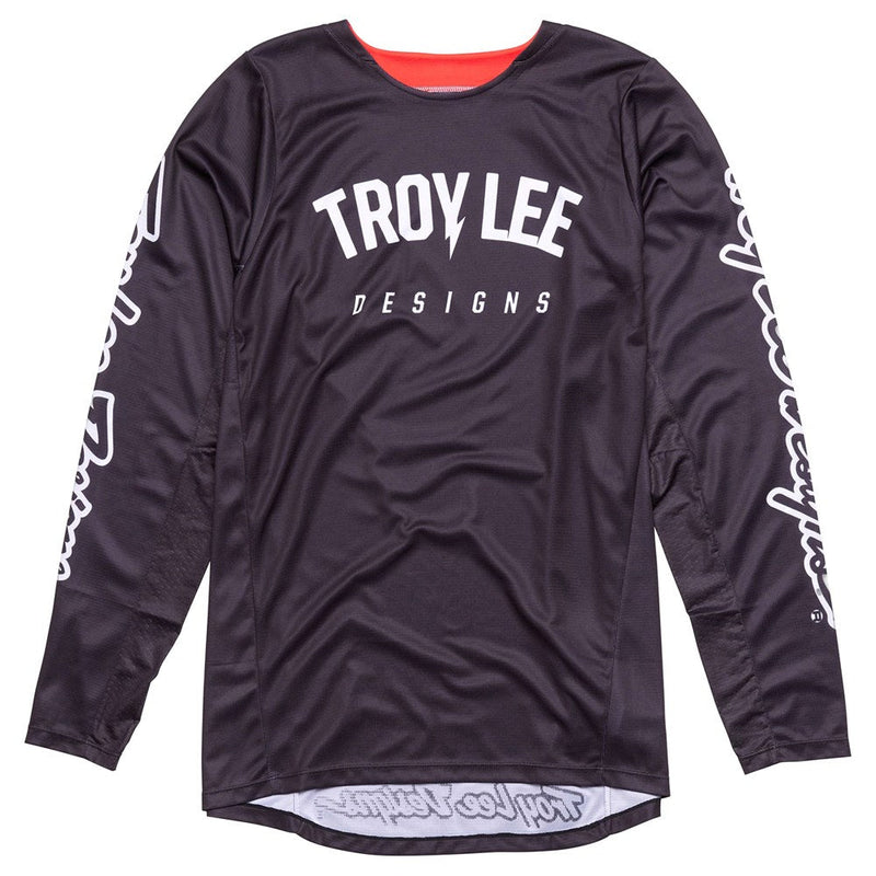 A person wearing the GP PRO JERSEY BOLTZ BLACK by Troy Lee Designs, featuring "TROY LEE DESIGNS" printed in white on the chest and sleeves, paired with red pants, red and black gloves, and a matching helmet. They are holding their hands out in front.