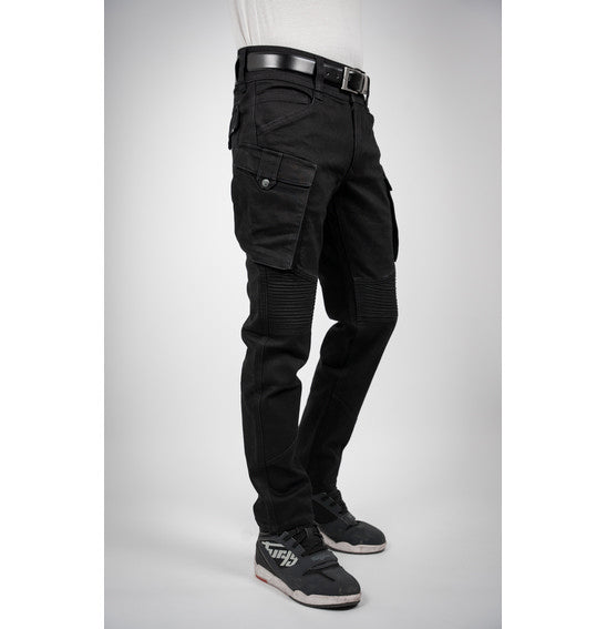 A person wearing Bull-It's jet black Jackal Motorcycle Jeans in a combat style, paired with a black belt and black sneakers with white soles, stands facing away. The jeans feature detailed stitching and zipper accents. The background is plain light gray.