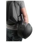 A person wearing a gray shirt and jeans carries a black motorcycle helmet in their right hand. Sporting an Ogio MACH 3 Motorcycle Backpack - Stealth, known for its water resistance, the person also has a removable helmet carry strap on their black and gray backpack. The background is plain white.