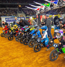 At a dirt track event, children lined up on STACYC 12eDRIVE electric balance bikes with 12” composite wheels, wearing colorful helmets and gear. A crowd watches in the background, and flags hang above them, capturing the vibrant energy of the scene.