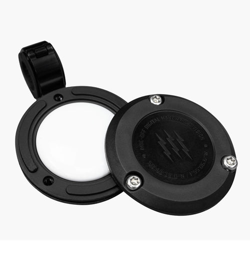 Close-up of two hands working with a circular, black mechanical component featuring a raised relief design. The hands are positioned near the terminals of the Muc-Off Secure Air-Tag Holder - Bike Security by Muc-Off, manipulating wires connected to it, similar to installing an Apple AirTag with anti-theft security screws. The background is dark and blurry.