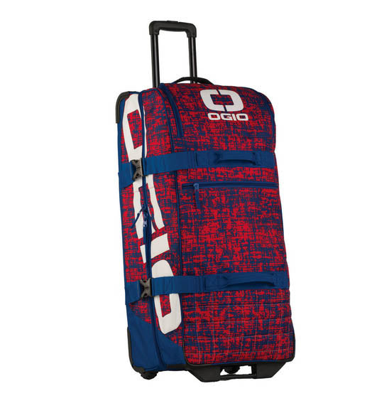 The OGIO TRUCKER Gear Bag in Chaos Red/Blue has an all-terrain wheel set, retractable handle, red and blue abstract pattern with white accents, and displays the OGIO logo on the front.