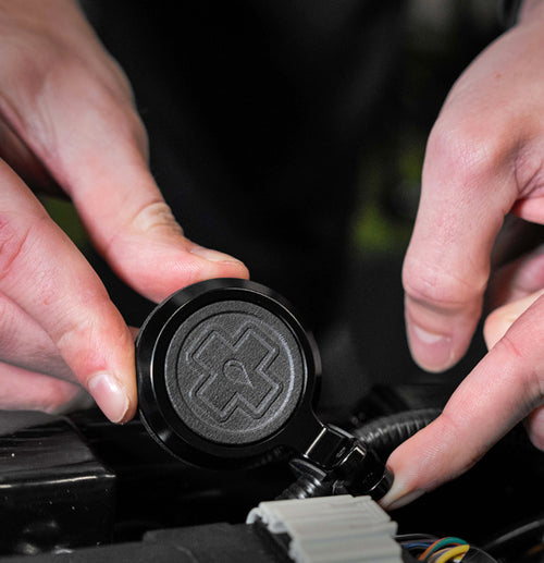 Close-up of two hands working with a circular, black mechanical component featuring a raised relief design. The hands are positioned near the terminals of the Muc-Off Secure Air-Tag Holder - Bike Security by Muc-Off, manipulating wires connected to it, similar to installing an Apple AirTag with anti-theft security screws. The background is dark and blurry.