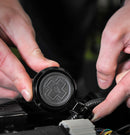 Close-up of two hands working with a circular, black mechanical component featuring a raised relief design. The hands are positioned near the terminals of the Muc-Off Secure Air-Tag Holder - Bike Security by Muc-Off, manipulating wires connected to it, similar to installing an Apple AirTag with anti-theft security screws. The background is dark and blurry.