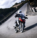 A motorcyclist wearing a white helmet and protective gear rides a red and white motorcycle fitted with METZELER KAROO STREET - Adventure (On/Off) tyres on a curving mountain road. The landscape in the background features trees and rugged terrain under a cloudy sky, emphasizing the bike’s impressive on-road performance.
