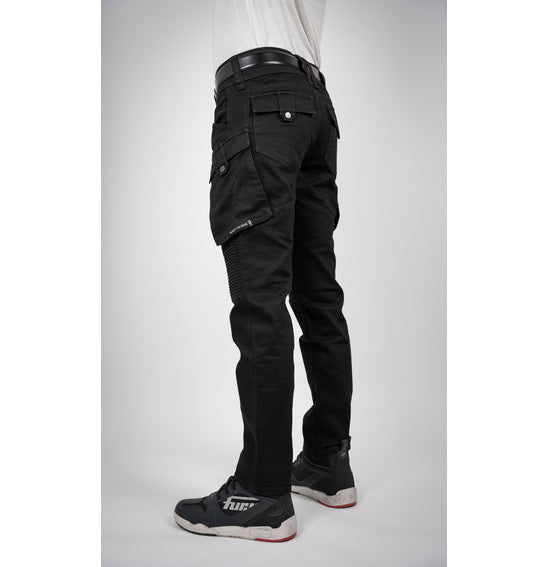 A person wearing Bull-It's jet black Jackal Motorcycle Jeans in a combat style, paired with a black belt and black sneakers with white soles, stands facing away. The jeans feature detailed stitching and zipper accents. The background is plain light gray.