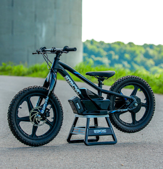A STACYC 18eDRIVE Brushless - Electric Balance Bike in black, featuring a sleek design, is displayed outdoors on a paved surface. It is powered by a 36 Volt BRUSHLESS motor and is resting on a stand with a lush, green landscape in the background.