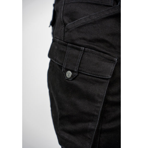 A person wearing Bull-It's jet black Jackal Motorcycle Jeans in a combat style, paired with a black belt and black sneakers with white soles, stands facing away. The jeans feature detailed stitching and zipper accents. The background is plain light gray.