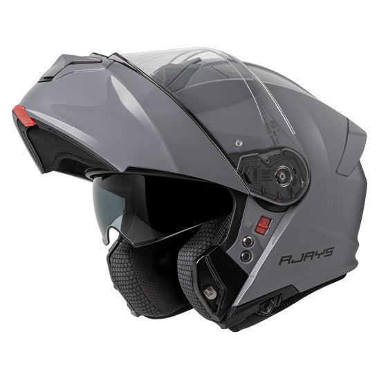 The RJAYS TOURTECH V Helmet in Solid Gloss Grey is a sleek, full-face motorcycle helmet featuring a flip-up front visor. Its aerodynamic shell includes an internal sun visor, a red button for visor release, and a cushioned interior lining. The brand name "RJAYS" is prominently displayed on the side.