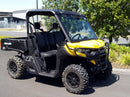 A Ventura Can-Am Defender HD5, 8, 10 (20 ->) in yellow with black accents and a robust towbar is parked on pavement near green bushes. Equipped with rugged tires for rough terrains, the utility vehicle stands ready. A building and some fencing can be seen in the background.