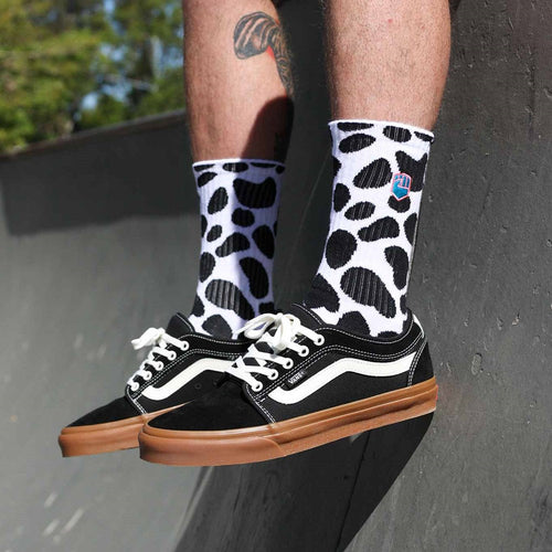 The DALMATION SOCK by FIST is a pair of white crew socks adorned with black irregular spot patterns reminiscent of cow print. These ultra-soft cotton socks also showcase a small colorful graphic near the top and offer targeted arch compression for enhanced support. They are shown against a white background.