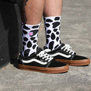 The DALMATION SOCK by FIST is a pair of white crew socks adorned with black irregular spot patterns reminiscent of cow print. These ultra-soft cotton socks also showcase a small colorful graphic near the top and offer targeted arch compression for enhanced support. They are shown against a white background.