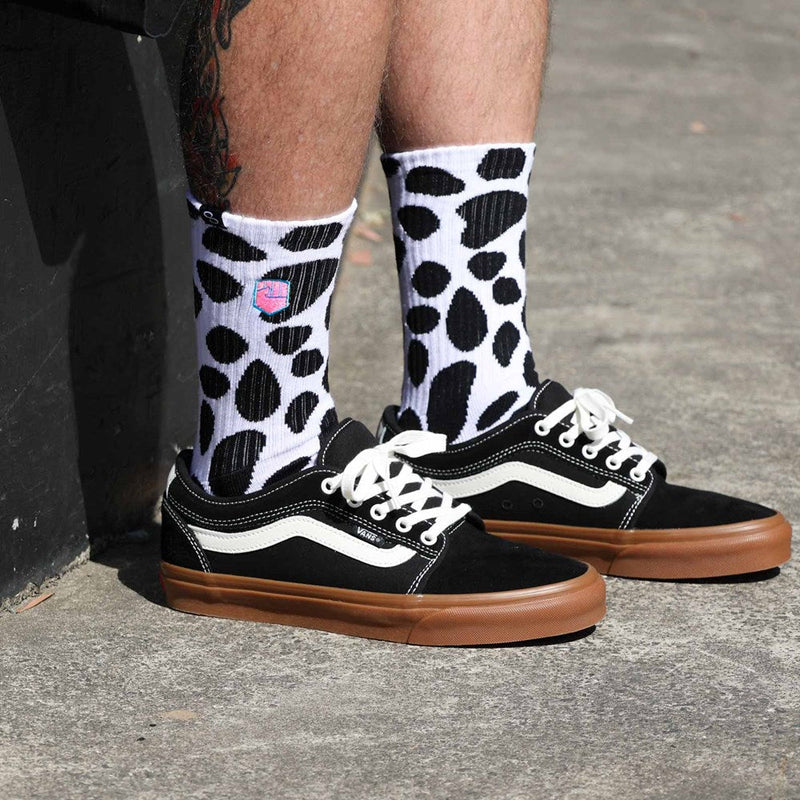 The DALMATION SOCK by FIST is a pair of white crew socks adorned with black irregular spot patterns reminiscent of cow print. These ultra-soft cotton socks also showcase a small colorful graphic near the top and offer targeted arch compression for enhanced support. They are shown against a white background.