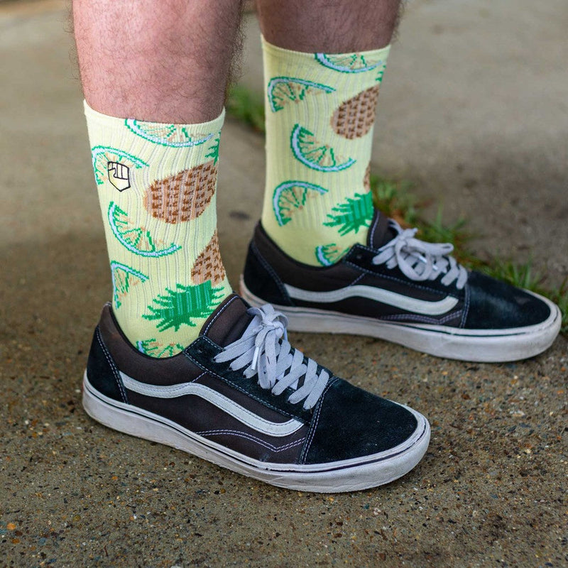 The PINA COLADA SOCK by FIST is a pair of beige crew socks adorned with a lively pattern of green pineapples and brown waffle cones, crafted from ultra-soft cotton.