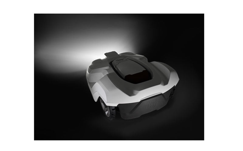 A Husqvarna robotic lawn mower with a sleek, futuristic design and compact body is shown from a rear angle. It is illuminated against a dark background, highlighting its contours and wheels. The robot features the Husqvarna Automower® Headlights Kit, suggesting its capability for autonomous navigation and lawn maintenance even at night.