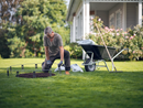 An older man kneels on backyard grass, tending a circular garden bed with soil. A Husqvarna Temporary Fence Kit and gardening supplies provide protection nearby, as blooming rose bushes line the house in the background.