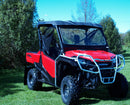 A red Honda ATV Pioneer SXS 1000, branded by Ventura, sits on the grass with a roof, protective bars, sturdy bull-bars, large tires, and black accents. Trees and a clear blue sky provide a picturesque backdrop.