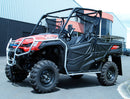 A Ventura Honda Pioneer 1000-5P in red and black, featuring roll bars and rugged tires, is parked on a paved area before a large industrial building with a corrugated metal facade.