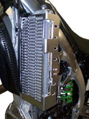 A close-up of a motorcycle radiator featuring the Emperor Racing Radiator Guard. It's encased in a red and white fairing, with visible engine and exhaust pipes below. Parts of the frame and a rear-view mirror are also seen.