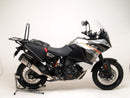 A blue adventure motorcycle, equipped with dual exhausts and off-road tires, is parked on a stand against a white background. It features a windshield and a black rear-mounted Tough-Rack by Ventura for luggage.