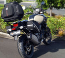 A blue adventure motorcycle, equipped with dual exhausts and off-road tires, is parked on a stand against a white background. It features a windshield and a black rear-mounted Tough-Rack by Ventura for luggage.